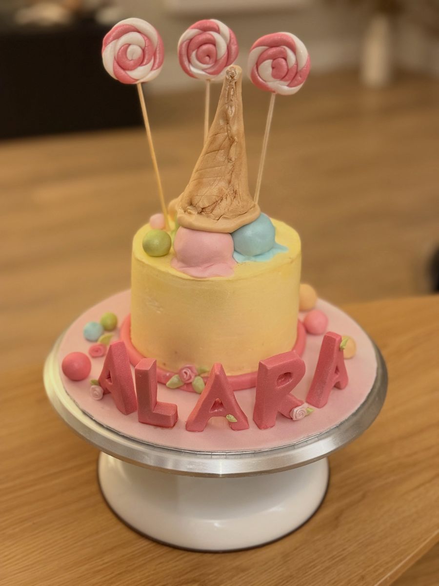 Two-tier pastel cake topped with handmade lollipops and the name Alara in pink fondant letters